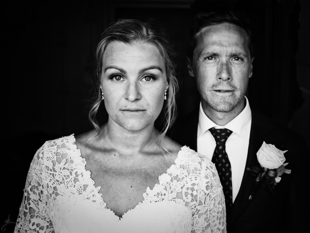 Black and white portrait of a bride and groom standing close together against a dark background, looking directly at the camera, with the bride in a lace dress and the groom in a suit with a boutonniere.