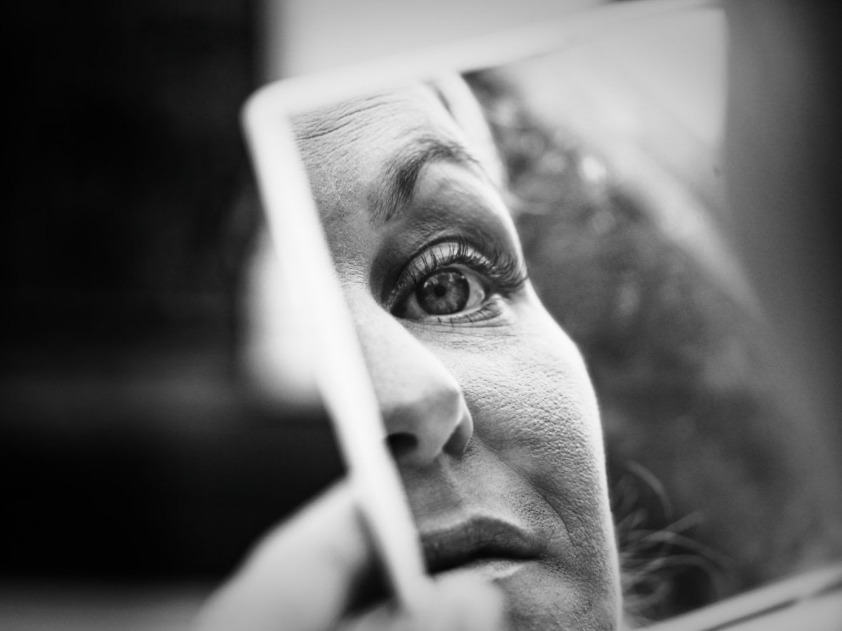 Black and white close-up photograph showing the reflection of a woman's eye and partial face in a small handheld mirror.