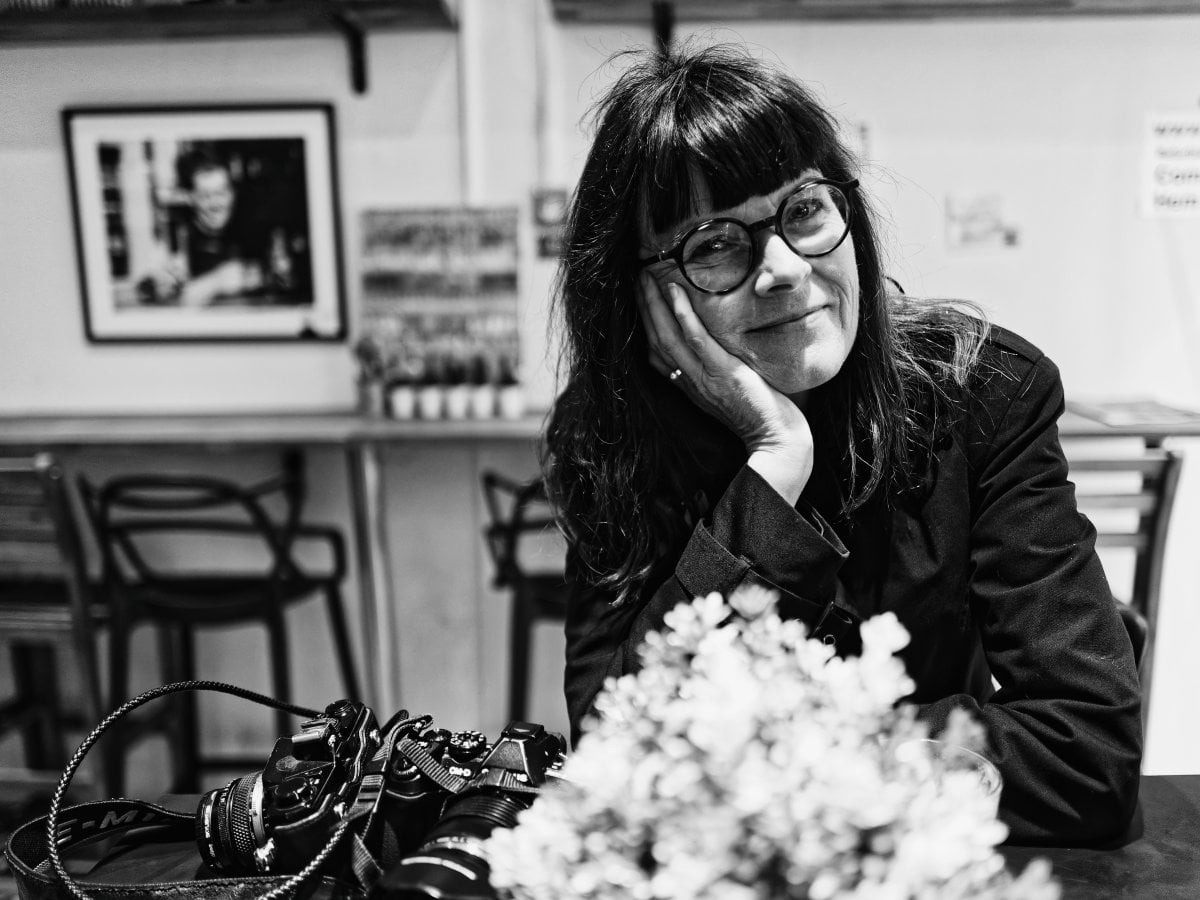 Black and white portrait of a smiling woman with bangs and glasses resting her cheek on her hand at a table, with a camera and blurred flowers in the foreground.