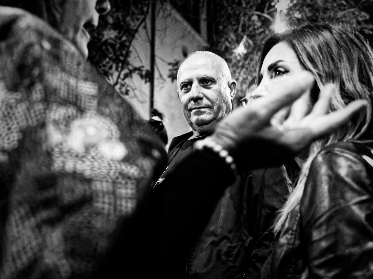 Black and white candid photograph of a bald man looking forward, framed by two blurred women in the foreground in a night setting.