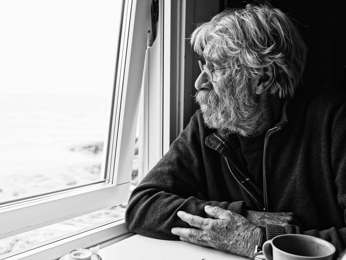 Black and white portrait of an elderly bearded man with glasses sitting at a table and looking out a window.