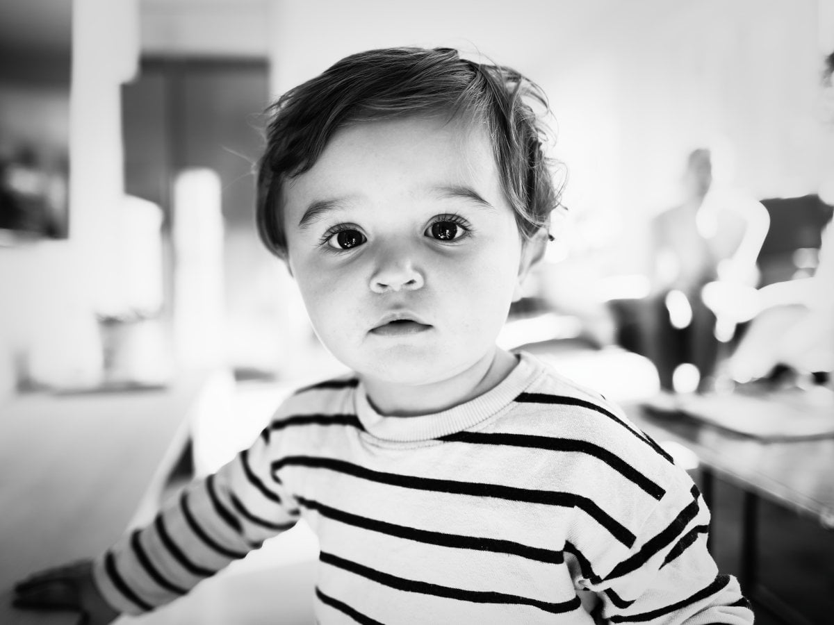 Black and white close-up portrait of a toddler wearing a striped long-sleeved shirt, looking directly at the camera with wide eyes against a blurred background.