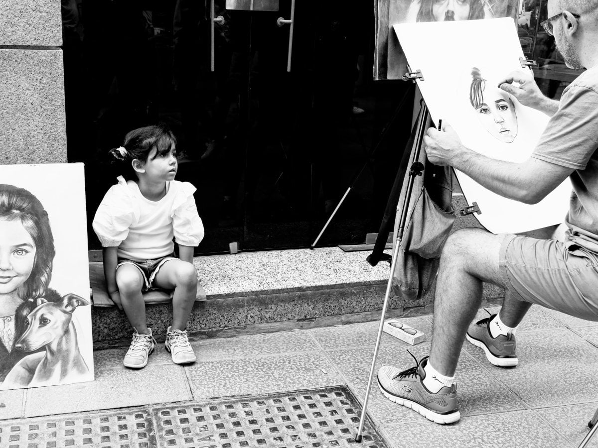 Black and white street photograph of an artist sketching a portrait of a young girl posing on a ledge, with a finished drawing leaning against the wall beside her.