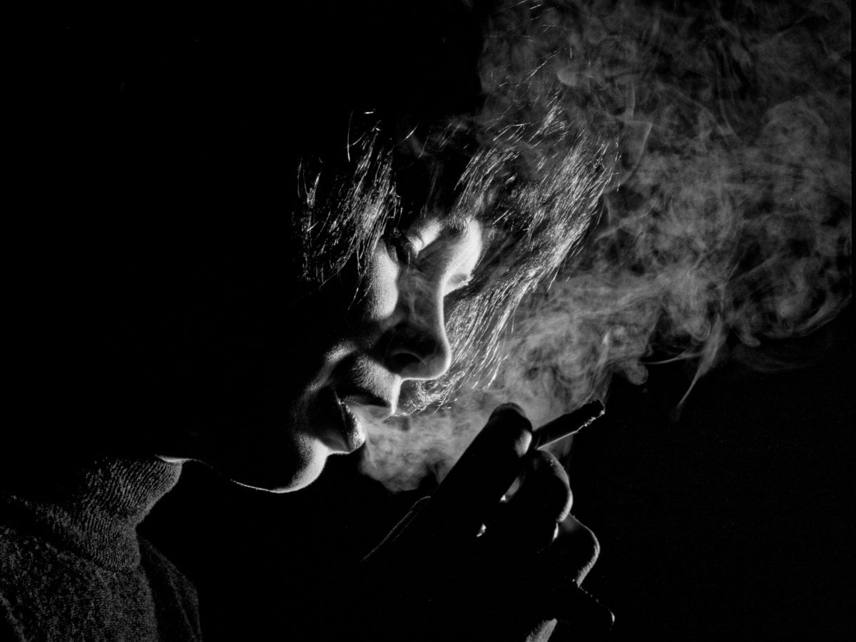 Black and white profile portrait of a person smoking, with dramatic backlighting highlighting the swirling smoke and their hair against a dark background.