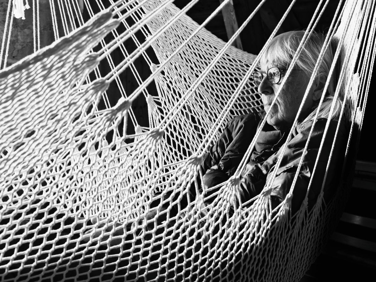 Black and white portrait of an older woman with glasses resting in a rope hammock, viewed through the hammock's netting.