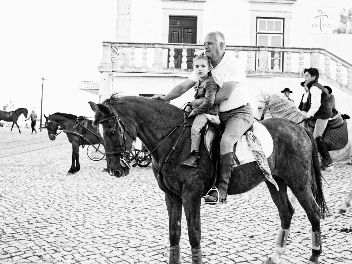 Black and white photograph of a man and a young girl riding together on a horse in a cobblestone square, with other horses and riders visible in the background.