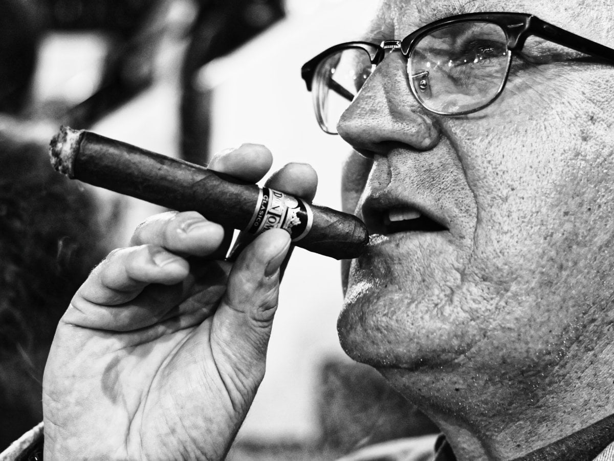 Black and white close-up portrait of a man wearing glasses, holding a large cigar near his mouth.