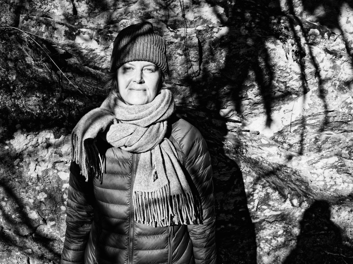 Black and white portrait of a woman wearing a beanie, scarf, and puffer jacket standing against a textured rock wall with dappled shadows.