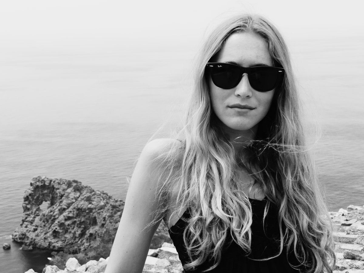 Black and white portrait of a woman with long wavy hair wearing sunglasses, standing against a backdrop of the sea and a rocky cliff.