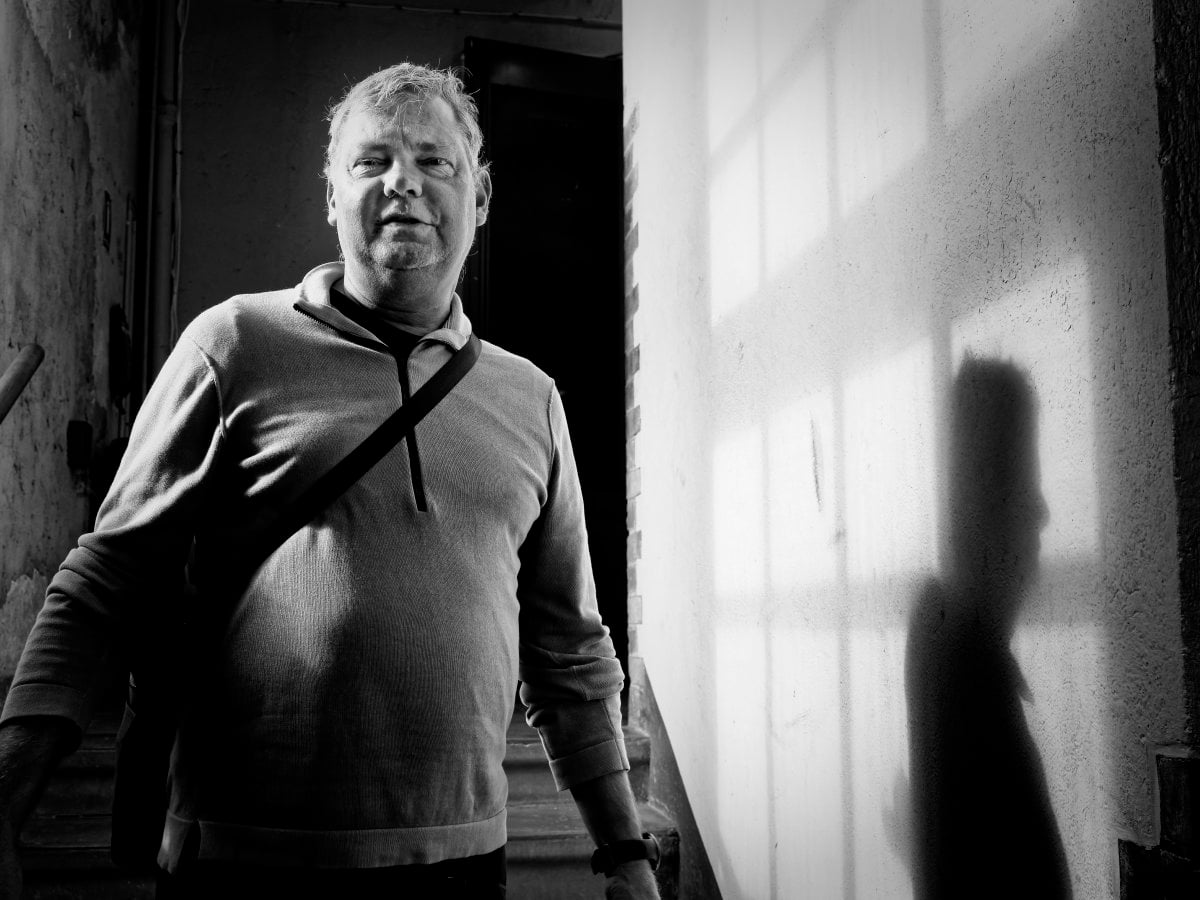 Black and white portrait of a man standing in a stairwell, wearing a half-zip sweater, with strong side lighting casting a sharp shadow of his profile and a window frame onto the wall.