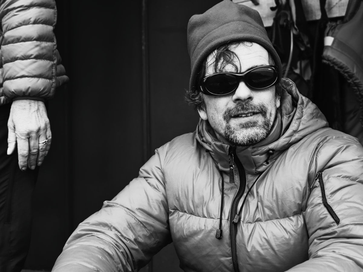 Black and white portrait of a bearded man wearing a beanie, sunglasses, and a puffer jacket, with the arm and hand of another person visible on the left edge.