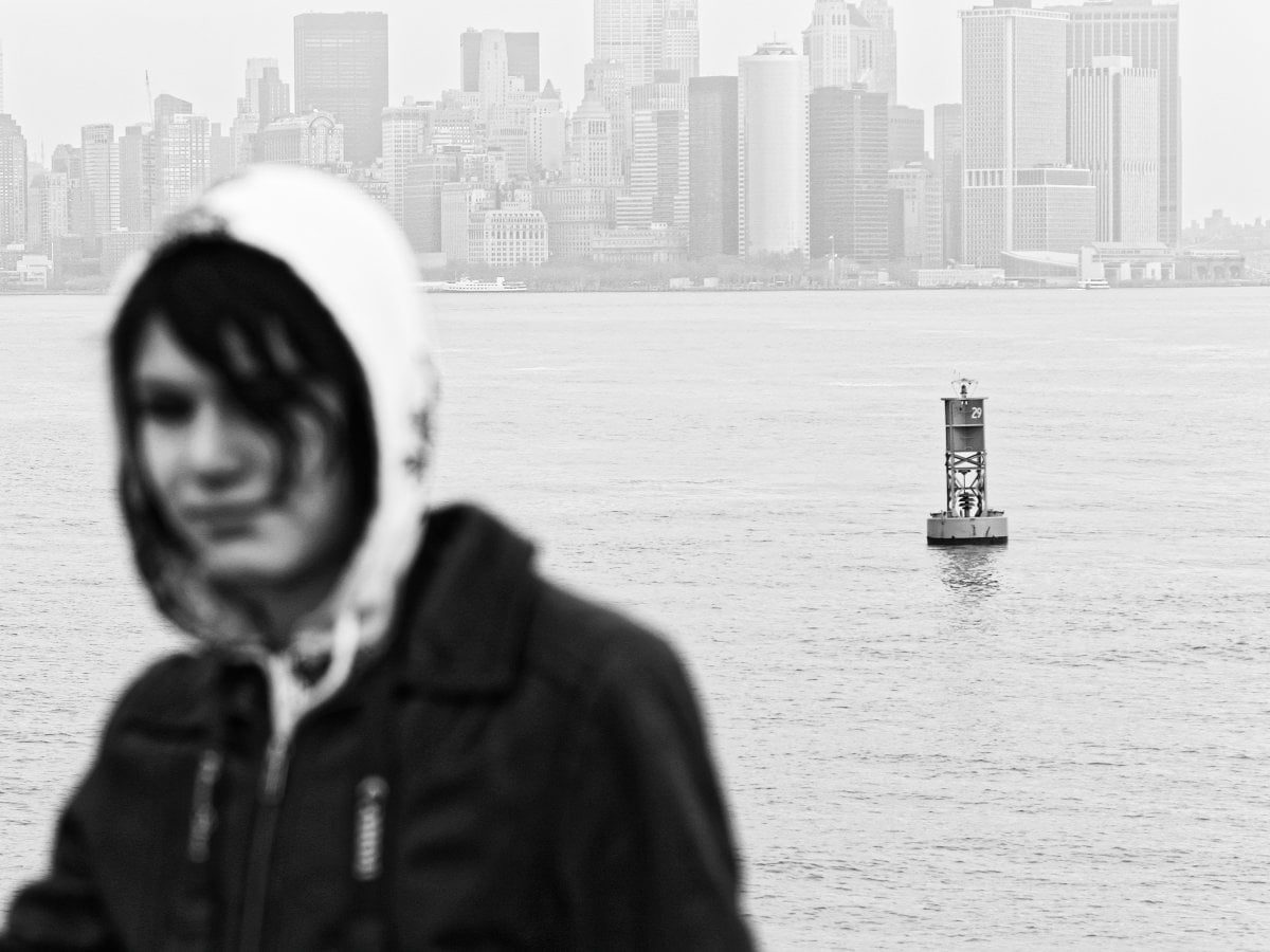 Black and white photograph focusing on a navigational buoy in the water, with a blurred person in a hooded jacket in the foreground and a city skyline in the background.