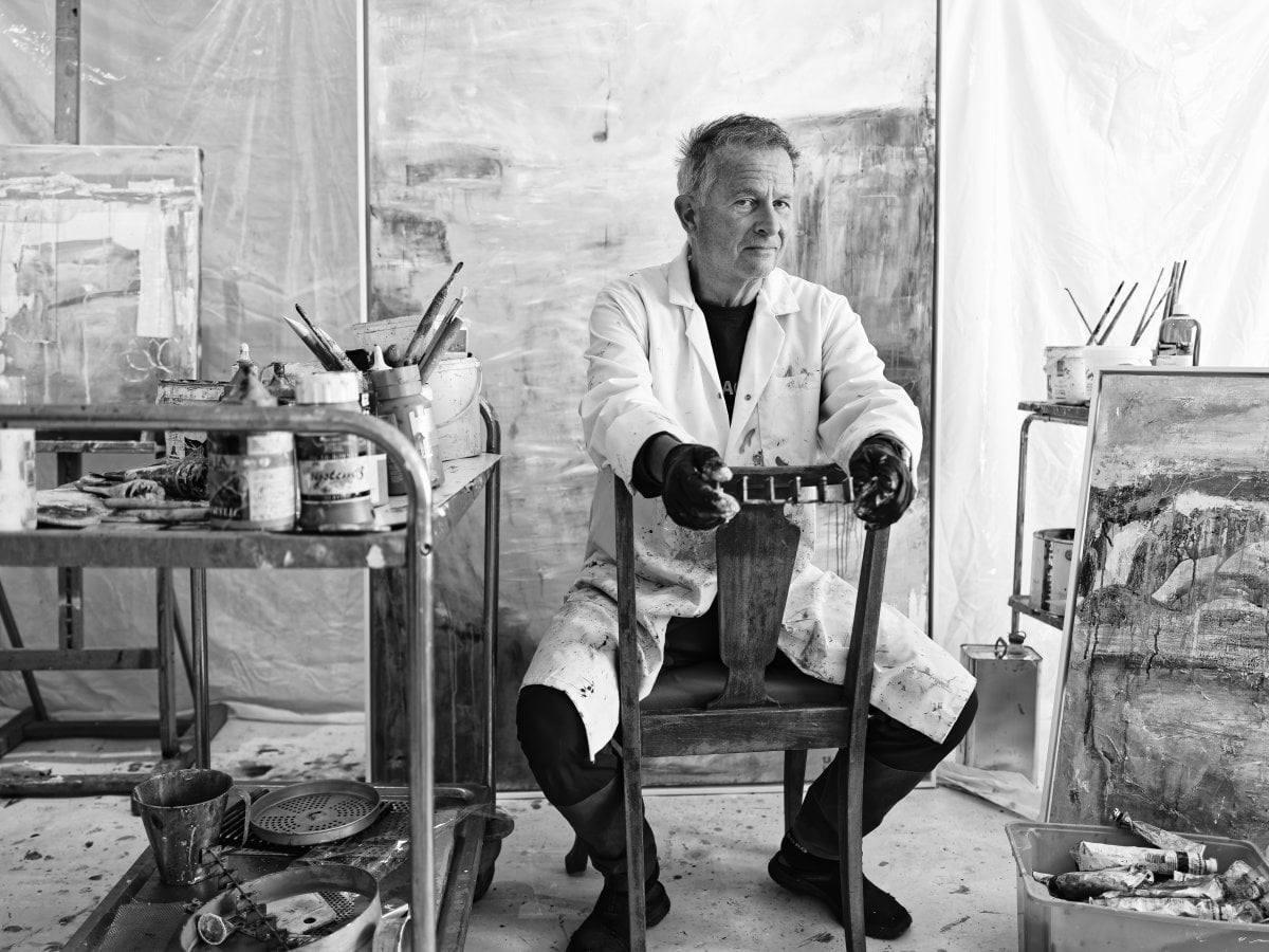 Black and white portrait of a male artist sitting backwards on a wooden chair in a studio, wearing a paint-splattered coat and gloves, surrounded by paints, brushes, and canvases.