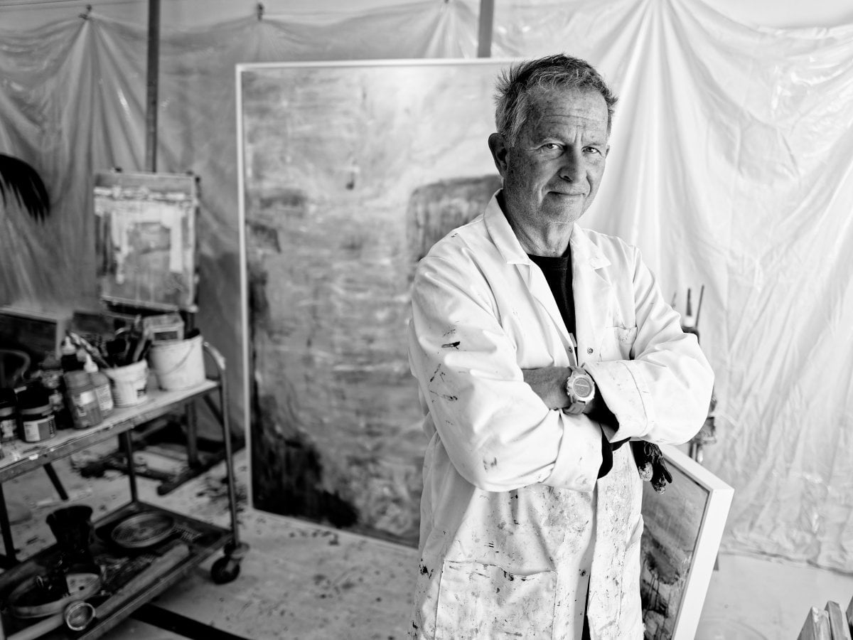 Black and white portrait of a male artist standing with crossed arms in a studio, wearing a paint-splattered white coat, with a large canvas and art supplies in the background.
