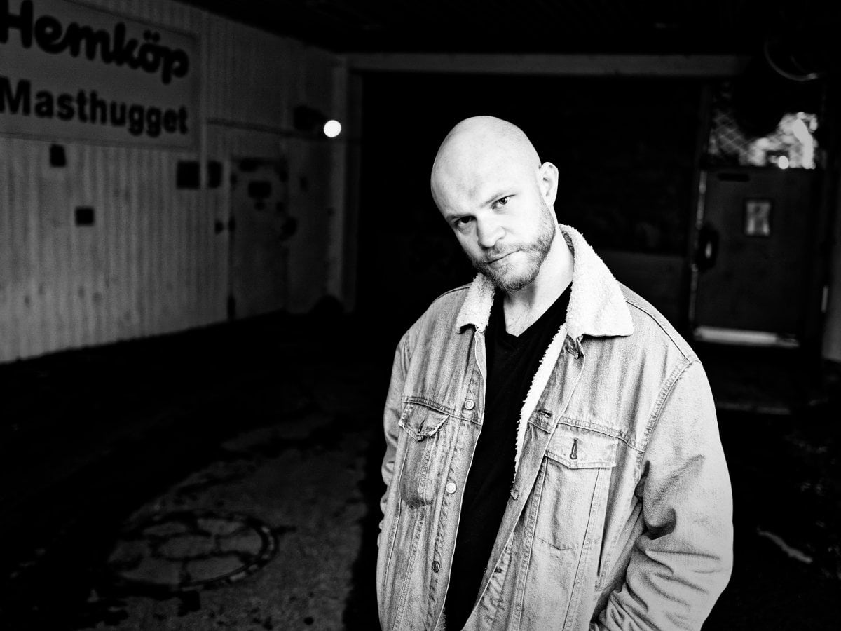 Black and white portrait of a bald man with a beard wearing a denim jacket with a shearling collar, standing in a dark urban environment with a sign reading 'Hemköp Masthugget' in the background.