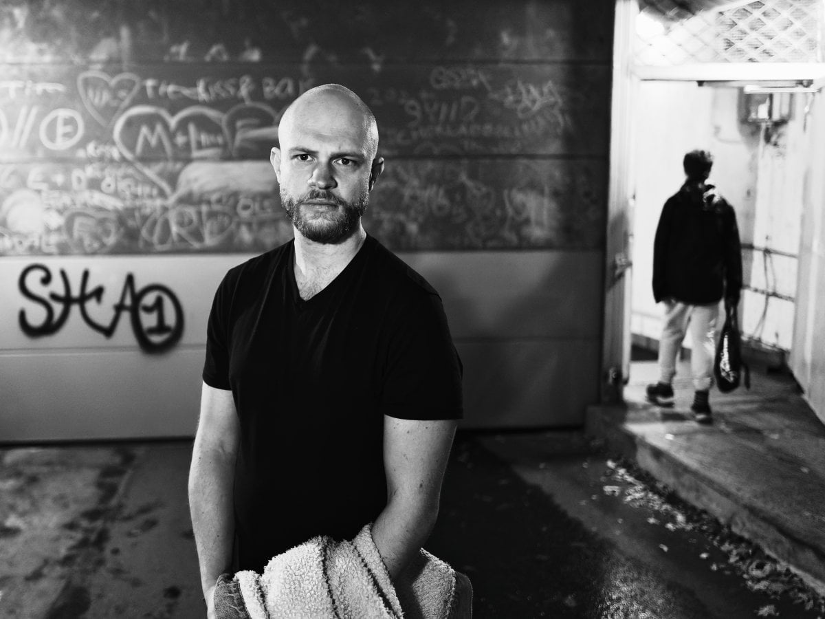 Black and white portrait of a bald man with a beard standing in front of a graffiti-covered wall, with a blurred figure walking away in the background.