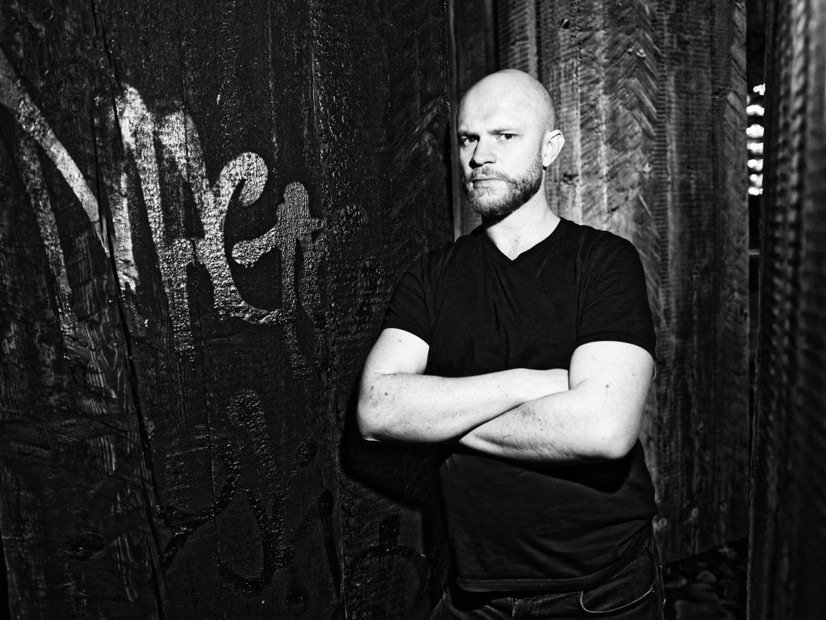 Black and white portrait of a bald man with a beard standing with crossed arms against a dark, textured wooden wall with graffiti.