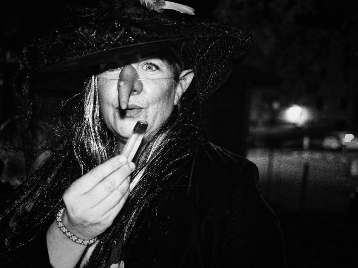 Black and white portrait of a woman in a witch costume with a large hat and fake nose, holding a cigarillo.